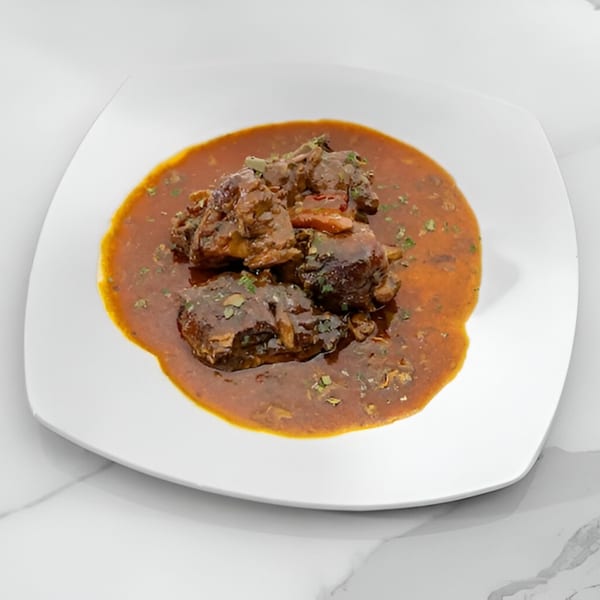 A white square plate holds Jamaican oxtail stew in a rich brown sauce, garnished with herbs.
