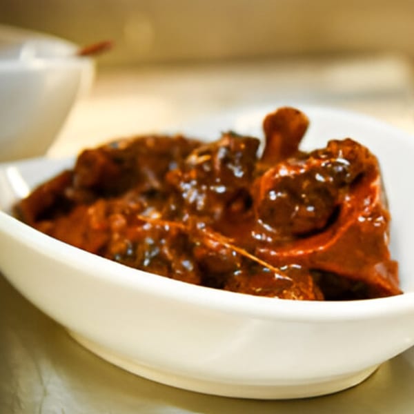 A white bowl filled with rich, dark brown Jamaican stewed chicken or goat.