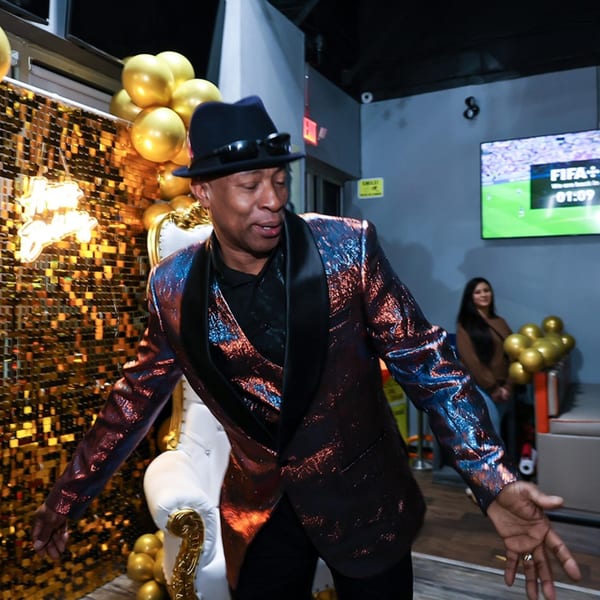 Man in a shiny suit and fedora dancing at Jewlz Bar, with golden decor and a TV.