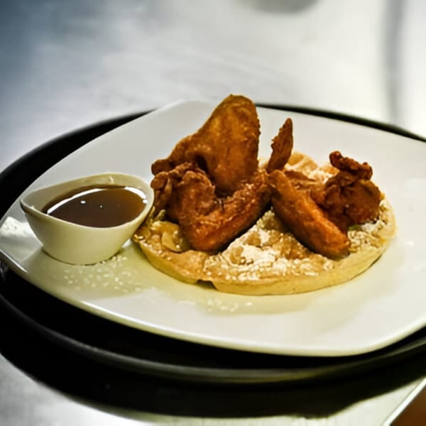Crispy fried chicken pieces on a waffle with powdered sugar, served with a side of syrup.